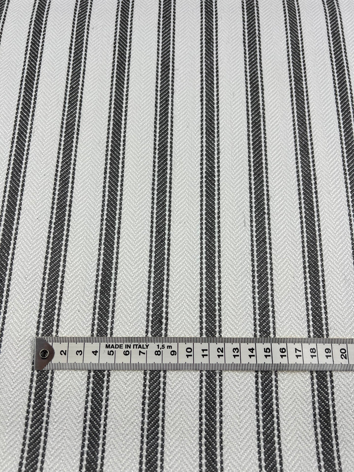 This upholstery fabric, Upholstery Herringbone - Tea Leaf - 145cm by Super Cheap Fabrics, features a black and white striped pattern, with evenly spaced vertical stripes. Highlighted by a measuring tape at the bottom, the scale is shown in centimeters. Part of our affordable range, this durable fabric is perfect for various decor projects.