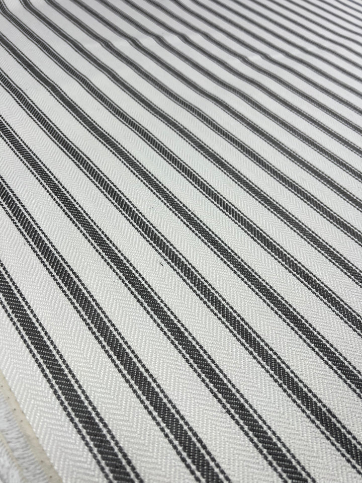 A close-up of Super Cheap Fabrics' Upholstery Herringbone - Tea Leaf - 145cm, an affordable upholstery fabric featuring a pattern of alternating thin, black and white stripes running diagonally. The durable fabric showcases a woven texture with slight variations in the stripe widths and a subtle sheen.