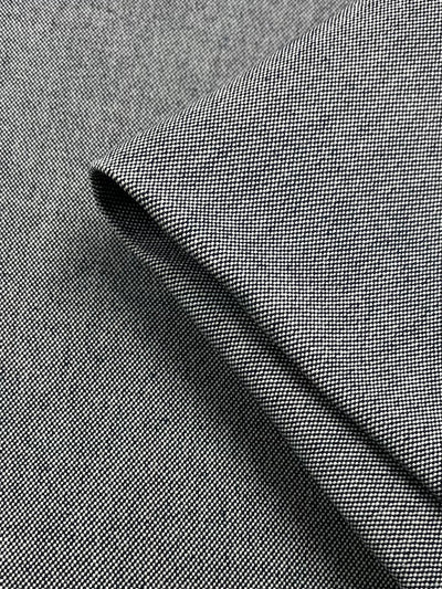 Close-up of a folded piece of black textured upholstery fabric. The image focuses on the intricate weave pattern, showcasing fine, tightly-knit fibers that give the heavyweight fabric its detailed and slightly rough appearance. One corner of the Super Cheap Fabrics Canvas - Black - 150cm is prominently folded over.