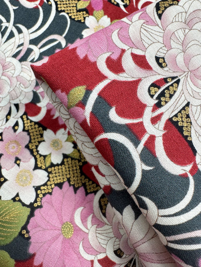 Close-up of Super Cheap Fabrics' Japanese Printed Cotton - Chrysanthemum (112cm), showcasing intricate pink, white, and red floral patterns with green leaves on a dark background, gold accents, and swirling traditional Japanese motifs.