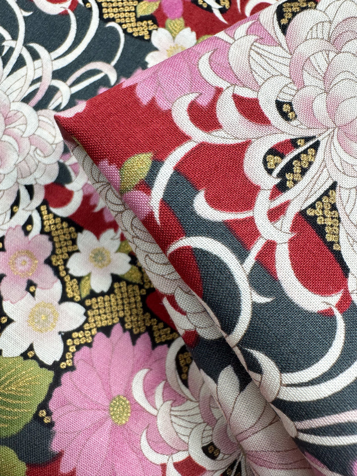 Close-up of Super Cheap Fabrics' Japanese Printed Cotton - Chrysanthemum (112cm), showcasing intricate pink, white, and red floral patterns with green leaves on a dark background, gold accents, and swirling traditional Japanese motifs.