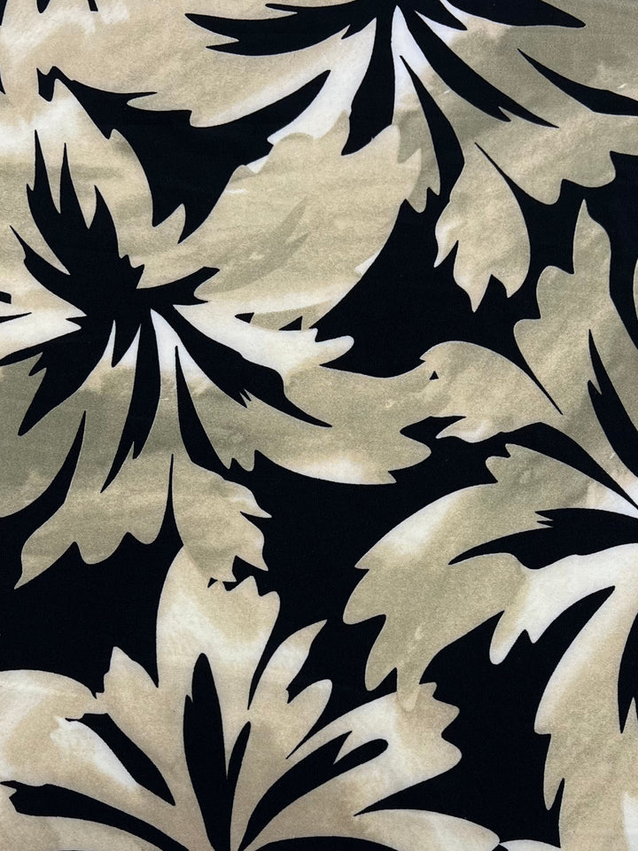 A close-up of a medium-weight fabric pattern featuring large, abstract leaf shapes in beige and light brown tones on a black background. The leaves are bold and stylized, creating a striking contrast against the dark backdrop, making this Super Cheap Fabrics Printed Lycra - Leafyworks - 150cm truly standout.