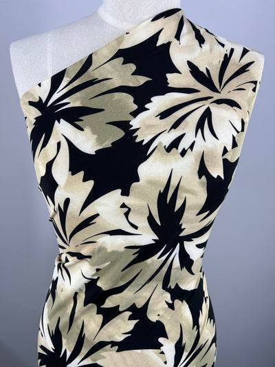 A dressmaker's mannequin displays a one-shoulder dress with a bold, abstract leaf pattern in Printed Lycra - Leafyworks - 150cm by Super Cheap Fabrics. The pattern features large, overlapping leaves in shades of beige, black, and cream. The neutral background ensures the medium-weight fabric of the dress is the focal point.
