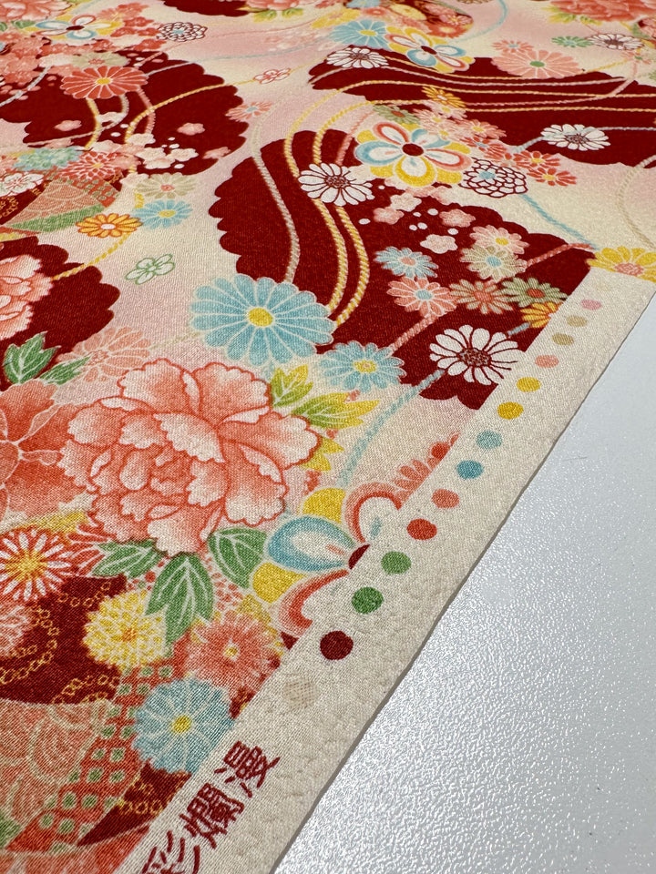 A close-up of Super Cheap Fabrics' Japanese Printed Cotton - Textured Crane Garden (112cm) shows vibrant red, pink, and yellow florals with green leaves, intricate motifs, colorful dots along the edge, and printed Japanese text.