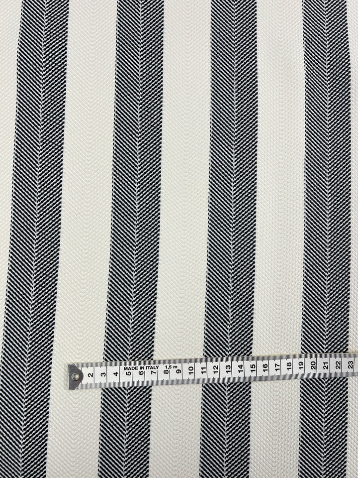 A close-up image of a **Super Cheap Fabrics Upholstery Herringbone - Salt & Pepper - 140cm** featuring vertical black and white chevron stripes. A measuring tape lies horizontally across the bottom, showing a length of 19 centimeters. The texture of the fabric appears fine and detailed, perfect for an affordable range of projects.