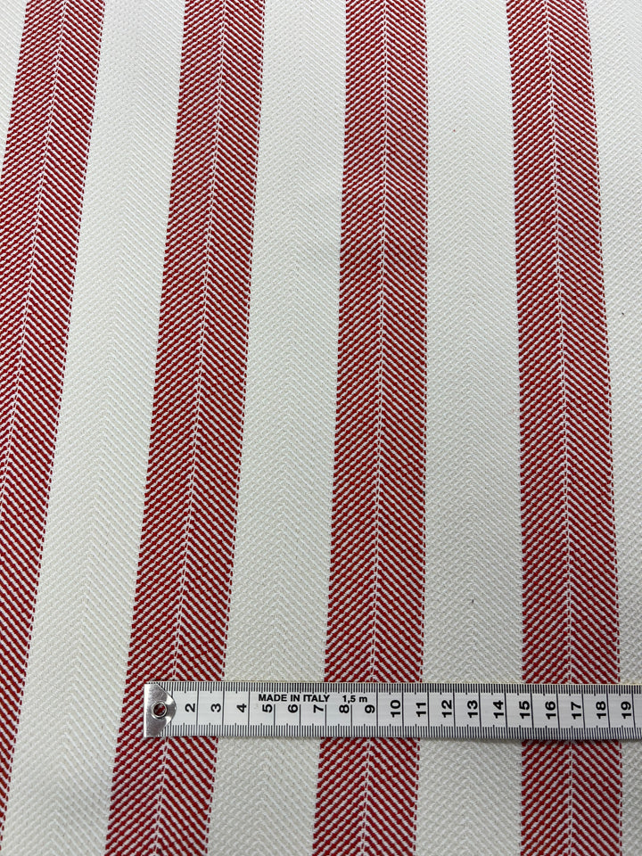A durable, stylish upholstery fabric with vertical red and white stripes, featuring a herringbone pattern within the red stripes. A white measuring tape with black numbers and markings is laid horizontally across the bottom portion of the fabric, showing measurements in centimeters. Product: Super Cheap Fabrics Upholstery Herringbone - Terracotta - 140cm