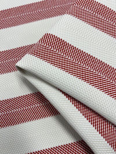 Close-up of an upholstery fabric with a patterned design in red and white. The red lines form a herringbone pattern, creating a diagonal striped effect against the white background. The durable fabric appears folded, showcasing its stylish appeal. This is the Upholstery Herringbone - Terracotta - 140cm from Super Cheap Fabrics.