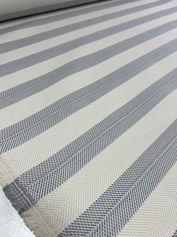A close-up view of an upholstery fabric roll displaying a striped pattern. The stripes alternate between light gray and off-white with a subtle herringbone texture, creating a sophisticated, elegant look. The material appears smooth and neatly woven, perfect for durable stylish fabrics that meet affordable upholstery needs. This is the Upholstery Herringbone - Silver - 140cm by Super Cheap Fabrics.