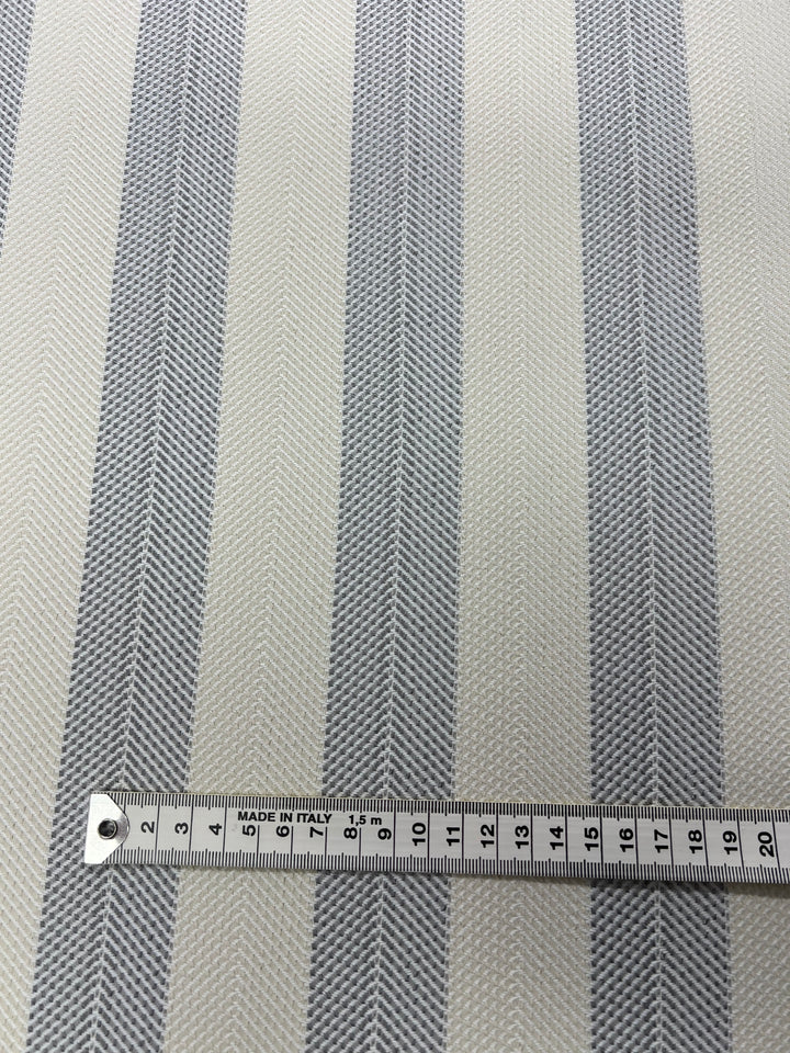 A close-up image of durable stylish upholstery fabric featuring vertical gray stripes spaced evenly on a light background. A measuring tape is placed horizontally at the bottom, marking the fabric's width in centimeters from 0 to 20. The measuring tape indicates "Upholstery Herringbone - Silver - 140cm" by Super Cheap Fabrics in black text.