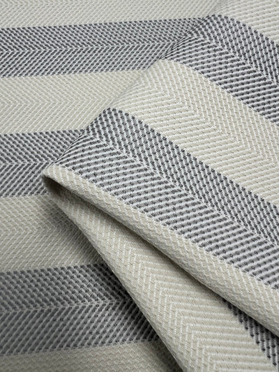 A close-up of folded Upholstery Herringbone - Silver - 140cm by Super Cheap Fabrics with a chevron pattern in alternating stripes of gray and off-white. The texture appears soft and woven, ideal for durable stylish fabrics, with the chevron stripes running diagonally across the fabric.