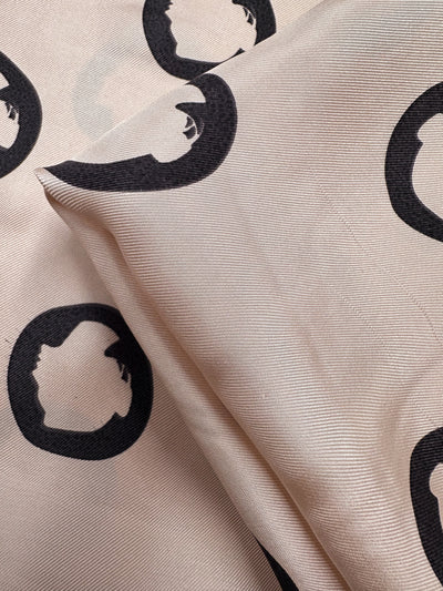 Super Cheap Fabrics' Pure Silk - Midnight Circles (140cm): Beige silk with black circular prints, each showing a stylized head profile. The luxurious fabric appears slightly wrinkled and is folded at the center.