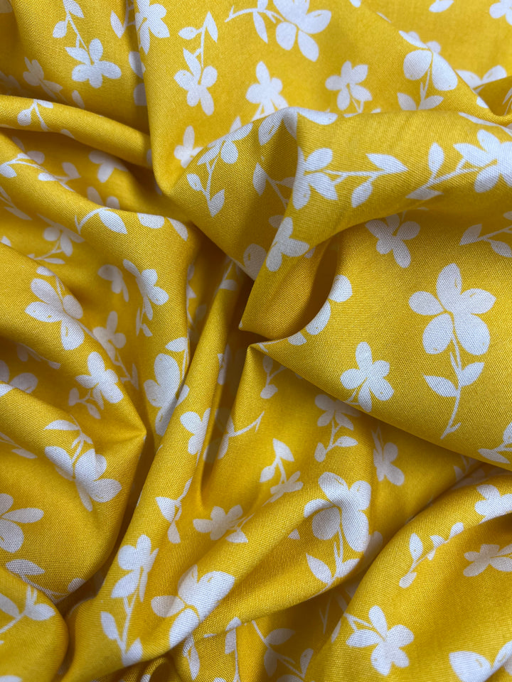 A close-up of a bright yellow **Printed Rayon - Yellow Jasmine - 145cm** fabric by **Super Cheap Fabrics** adorned with a white floral pattern. The material appears slightly crumpled, showcasing the vibrant color and delicate design of the flowers.