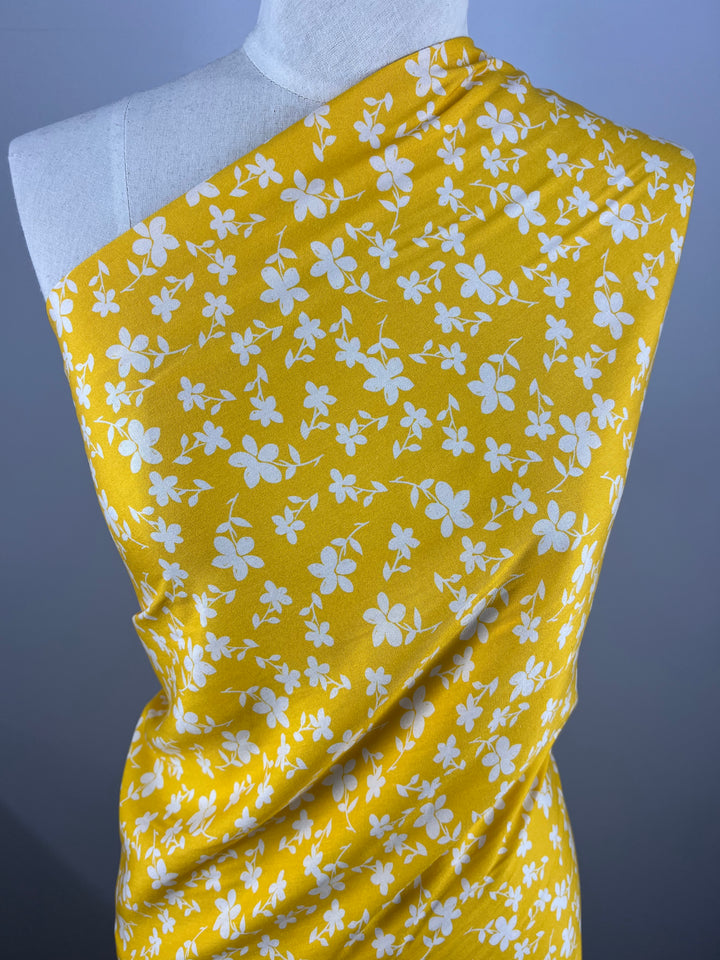 A yellow printed rayon fabric with white floral patterns draped over a white mannequin. The colourful fabric, Printed Rayon - Yellow Jasmine - 145cm by Super Cheap Fabrics, is arranged to cover one shoulder and elegantly flows down the front, showcasing the vibrant prints against a gray background.