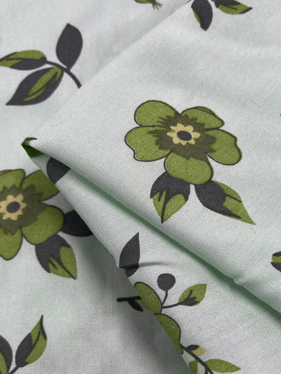 A close-up of a light green, lightweight fabric with a floral pattern. The design features green flowers with dark centers and dark green leaves, creating a fresh and natural look. The Super Cheap Fabrics' Printed Rayon - Green Dais - 145cm is gently folded, displaying the vibrant prints clearly.