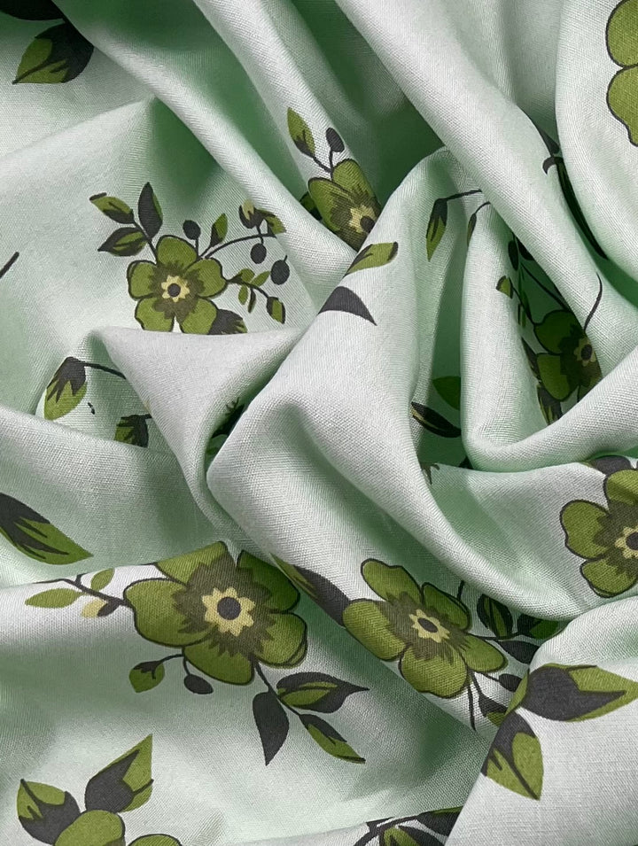 A close-up image of light green Printed Rayon - Green Dais - 145cm by Super Cheap Fabrics with a floral pattern. The lightweight fabric is crumpled, showcasing various shades of green flowers and leaves. The texture and folds of the material are prominently visible, highlighting its vibrant prints.