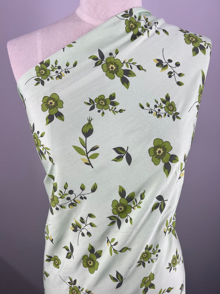 A mannequin is draped with Super Cheap Fabrics' Printed Rayon - Green Dais - 145cm, a lightweight mint green fabric featuring vibrant prints of dark green and yellow flowers, along with green leaves and stems. The background is plain and lavender in color.