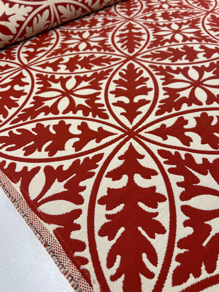 A close-up of the Upholstery Jacquard - Rooibos Tea - 144cm by Super Cheap Fabrics featuring red and white fabric with an intricate, symmetrical decorative pattern. The design consists of large red leaf-like shapes and interlocking motifs against a white background, creating a bold and elegant visual effect that's perfect for stylish, durable furnishings.