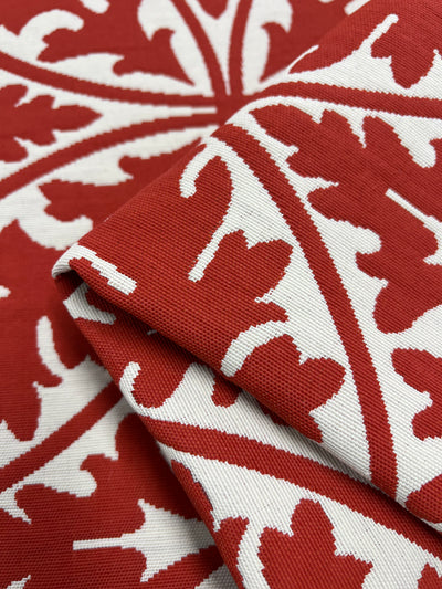 A close-up view of folded Upholstery Jacquard - Rooibos Tea - 144cm fabric by Super Cheap Fabrics featuring a red and white leafy pattern. The design consists of intricate red leaves on a white background, forming symmetrical, radial shapes. This stylish upholstery fabric's texture and detailed pattern are prominently displayed, showcasing its durability.