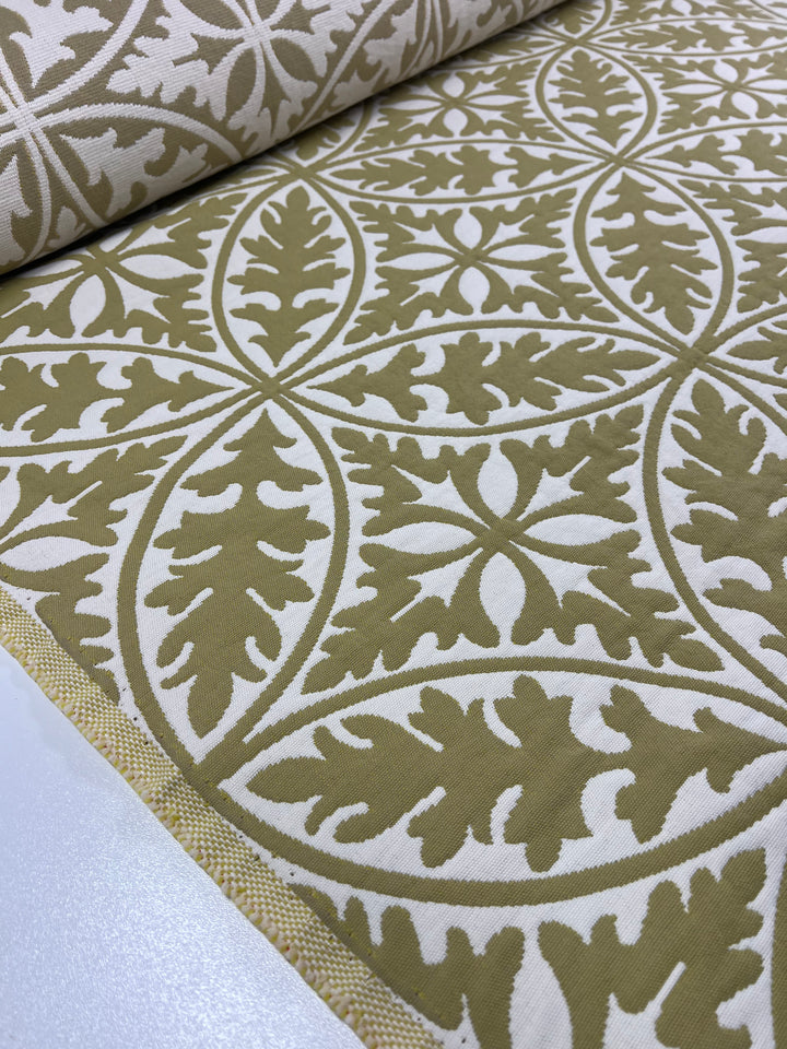 A close-up of Super Cheap Fabrics' Upholstery Jacquard - Antique Gold - 144cm with an intricate olive green and white pattern featuring symmetrical leaf-like designs. The durable fabric is partially unrolled on a flat surface, showcasing the continuous, repeating motif, part of our affordable range.