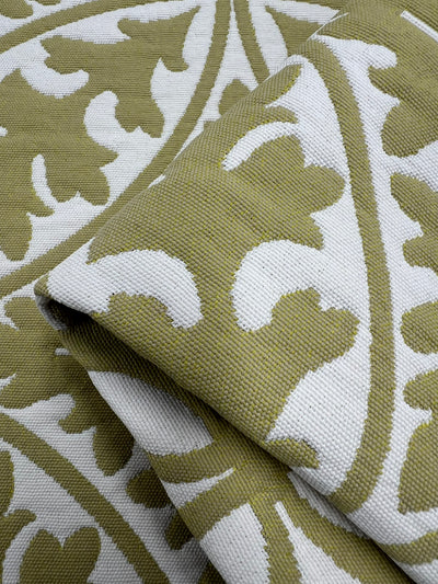 A close-up image of a folded Upholstery Jacquard - Antique Gold - 144cm with a green and white leaf pattern. The design features large, abstract leaf shapes arranged in a repeating pattern. The texture of the durable fabric is visible, highlighting its woven structure from an affordable range by Super Cheap Fabrics.