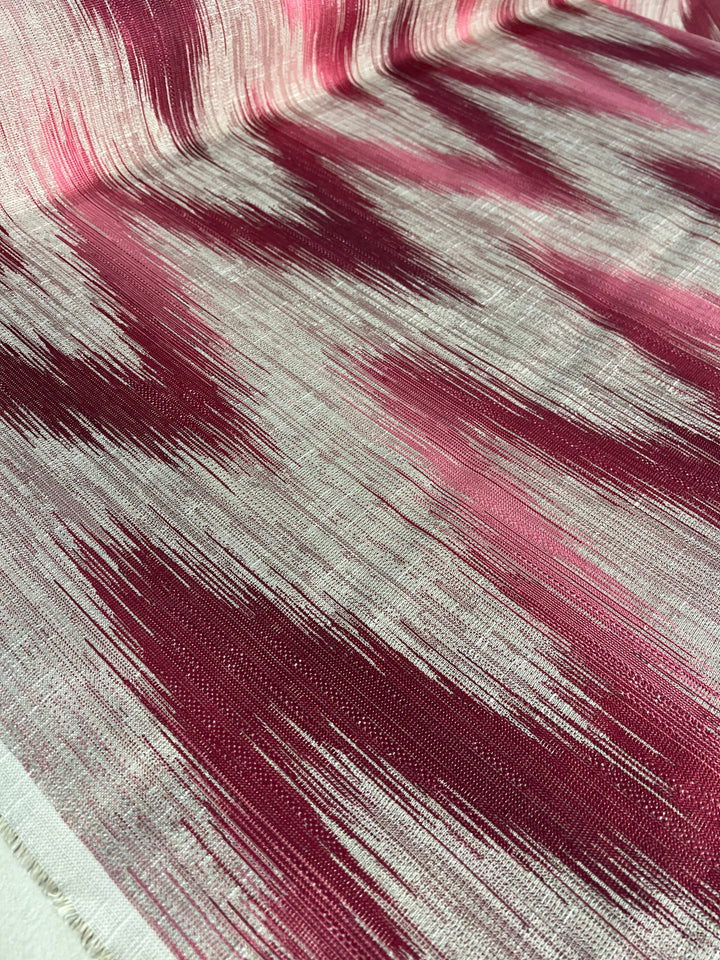 A close-up of Upholstery Jacquard - Beardsley Flames - 145cm by Super Cheap Fabrics, a durable upholstery fabric with an abstract pattern consisting of streaks of red, maroon, and pink on a beige background. The design resembles elongated brush strokes, creating a dynamic, flowing effect across the material's surface. Perfect for those seeking an affordable range without compromising on style.