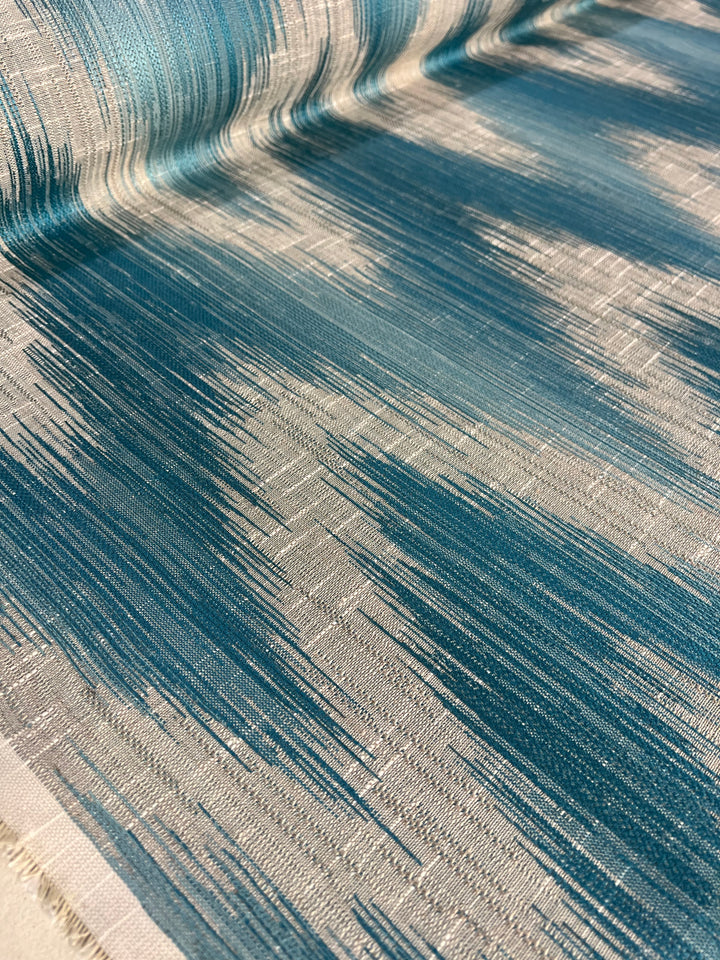 A close-up of the Upholstery Jacquard - Aqua - 145cm by Super Cheap Fabrics with a gradient pattern. The fabric features dynamic streaks of teal and silver colors, blending together to create a fluid, almost waterfall-like appearance. The texture of the durable fabric is visible, highlighting the intricacy of the weave.