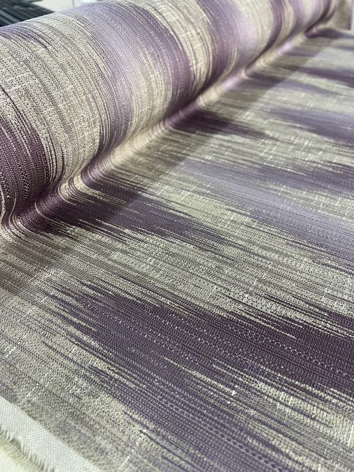 Close-up of a textured upholstery fabric with horizontal striations in shades of purple, silver, and beige. The material, slightly curled at the top edge, showcases its woven quality and gradient color transitions from our affordable range of durable stylish fabrics. This is the Upholstery Jacquard - Dusk - 145cm by Super Cheap Fabrics.