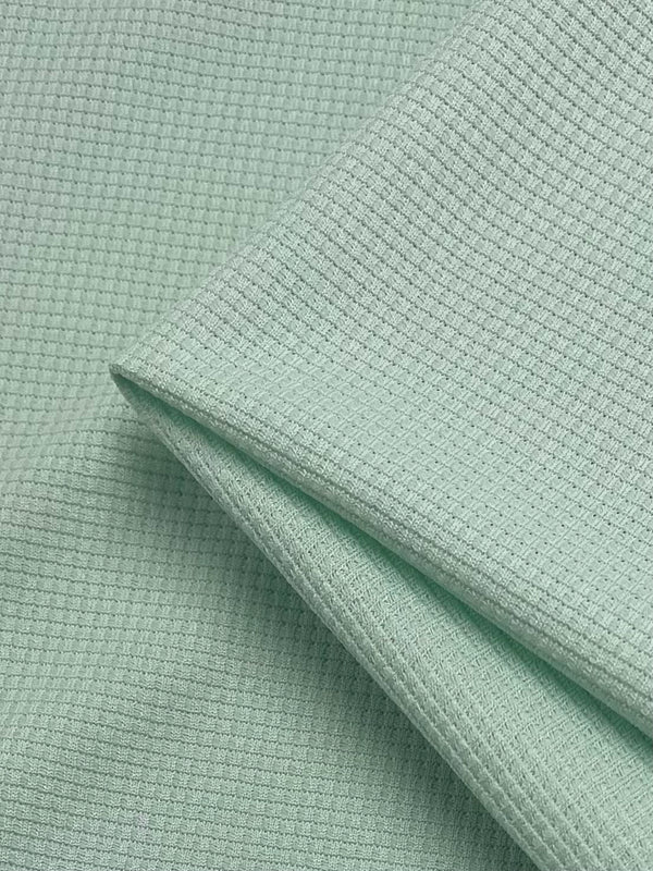 Close-up of a light green, textured waffle fabric with a small waffle pattern. The material is neatly folded to form clean edges, highlighting the intricate texture and soft appearance. The overall look is fresh and uniform. This is the Waffle Knit - Ambrosia - 170cm by Super Cheap Fabrics.