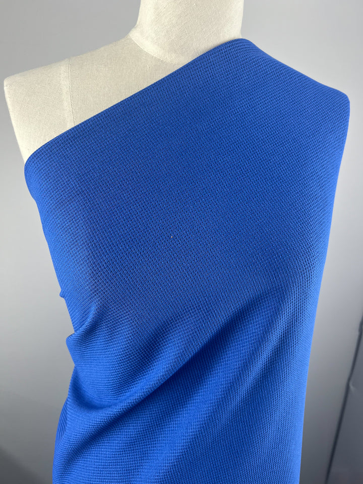 A mannequin is draped in a bright blue, Waffle Knit - Royal - 170cm from Super Cheap Fabrics that covers one shoulder. The medium-thickness material is tightly wrapped around the torso, creating a smooth and slightly structured appearance with a subtle three-dimensional texture. The background is a neutral gray.