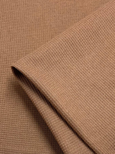 Close-up image of a folded piece of Waffle Knit - Mocha Mousse - 170cm by Super Cheap Fabrics. The texture of the material is visible, showcasing its fine, grid-like pattern and three-dimensional effect. One edge of the fabric is neatly folded over, creating a shadow that highlights the depth of the weave.