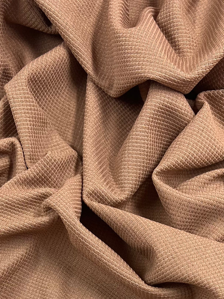 A close-up image of a textured beige waffle fabric with a slightly wrinkled surface. The fabric has a grid-like pattern, and folds create shadows and highlights, giving it a three-dimensional effect and crumpled appearance. The product featured is Waffle Knit - Mocha Mousse - 170cm by Super Cheap Fabrics.
