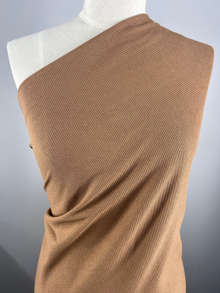 A close-up photograph of a Super Cheap Fabrics Waffle Knit - Mocha Mousse - 170cm draped around a mannequin. The fabric features a fine ribbed texture and wraps over one shoulder, creating smooth folds and a sophisticated appearance. The background is a plain gradient from light to dark gray.