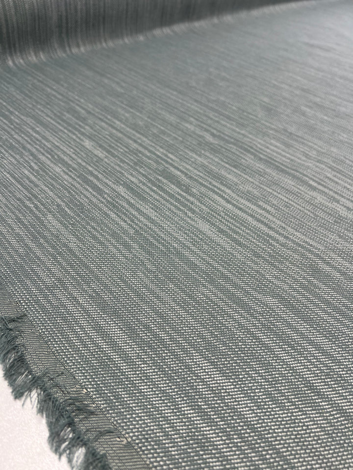 A close-up image of Super Cheap Fabrics' Upholstery Twill - Sea Mist - 147cm with a textured surface. The durable fabric appears smooth with fine horizontal lines and slightly frayed edges at one side, indicating it has been cut from a larger piece. The twill weave fabric is laid flat on a white surface.