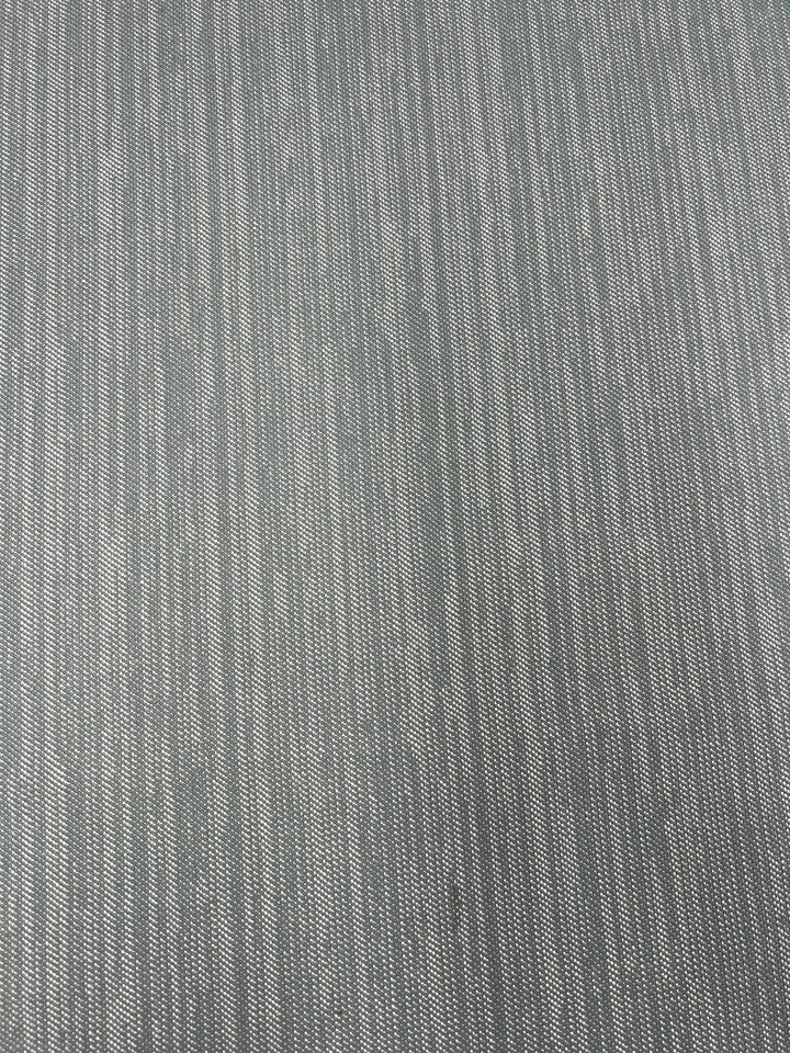 A close-up image of a textured gray fabric. The Upholstery Twill - Sea Mist - 147cm by Super Cheap Fabrics features a subtle pattern of vertical lines, varying in shades of gray and white, creating a slightly striped appearance. The durable fabric appears woven with a soft, uniform texture.