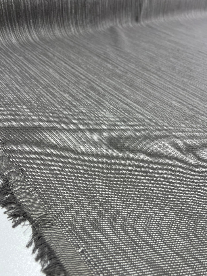 Image of a close-up view of a piece of Upholstery Twill - Dove Grey - 147cm by Super Cheap Fabrics with a fine, horizontal weave pattern. The durable fabric appears to be grey with a smooth texture. The edges are slightly frayed, and the weave is tight and uniform. Perfect for furniture upholstery, the fabric has a subtle shimmer.
