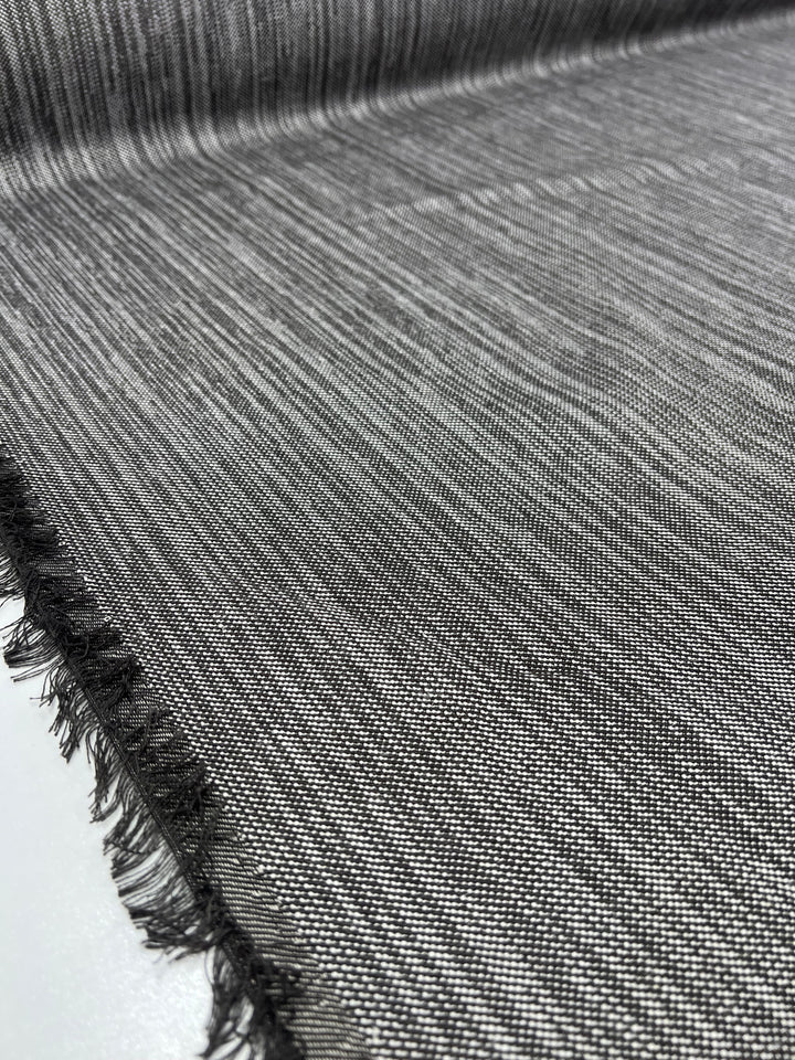 Close-up of a grey, finely woven **Super Cheap Fabrics Upholstery Twill - Salt & Pepper - 147cm** laid flat, featuring a subtle gradient from dark grey at the bottom edge to lighter towards the top. The bottom edge is frayed, revealing fringed threads. This durable fabric's texture is visibly tight and detailed, ideal for furniture upholstery.