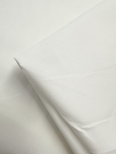 A close-up of Super Cheap Fabrics' Plain Cotton - Ivory - 150cm shows the 100% cotton fabric folded on a white surface, highlighting its subtle creases and soft shadows.