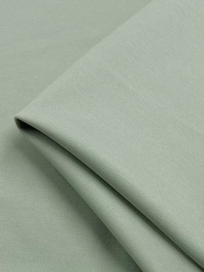 A close-up image of folded light gray-green fabric, displaying a smooth and slightly textured surface. The Super Cheap Fabrics Ponte - Desert Sage - 153cm is neatly arranged, showing clean lines and soft folds.