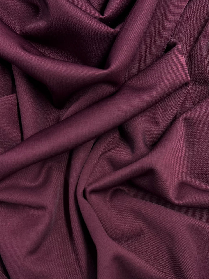 A close-up image of burgundy stretch fabric, showing its smooth and slightly shiny texture. The fabric is loosely folded and draped, creating soft shadows and highlights that emphasize the material's gentle, flowing nature. This is "Ponte - Cordovan - 153cm" from Super Cheap Fabrics.