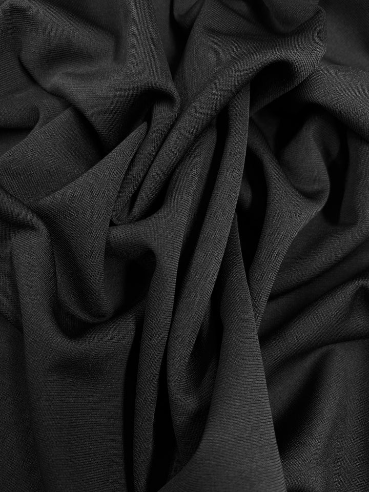 Super Cheap Fabrics' Ponte - Black - 150cm intricately folded and draped, creating soft, elegant curves and shadows throughout the texture. The mid-weight knit material appears smooth with a slightly matte finish and is wonderfully wrinkle-resistant.