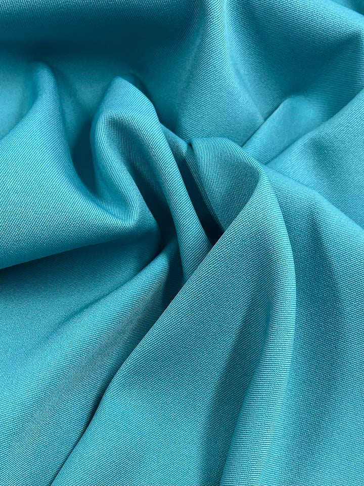 A close-up of crumpled, turquoise Twill Suiting - River Blue - 155cm fabric material from Super Cheap Fabrics creating soft folds and shadows. The diagonal weave pattern appears smooth and slightly shiny, capturing subtle variations in light and shade.