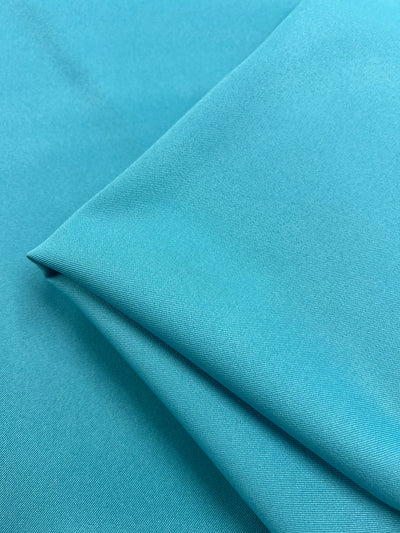 A close-up view of a neatly folded, Twill Suiting - River Blue - 155cm from Super Cheap Fabrics reveals its smooth, soft texture with a subtle sheen. The diagonal weave pattern of the 100% polyester material creates clean lines at the edges of the folds, emphasizing its pliable nature.
