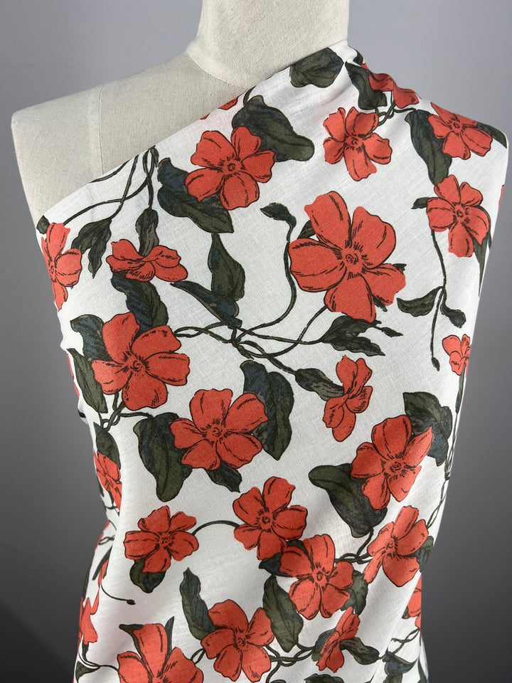 A mannequin draped in Super Cheap Fabrics' Printed Linen - Periwinkle - 135cm adorned with a vibrant floral print. The fabric features large red flowers with green leaves on a white background, arranged in a lively and elegant pattern. Perfect for garment creations, the backdrop is a simple, plain grey surface.