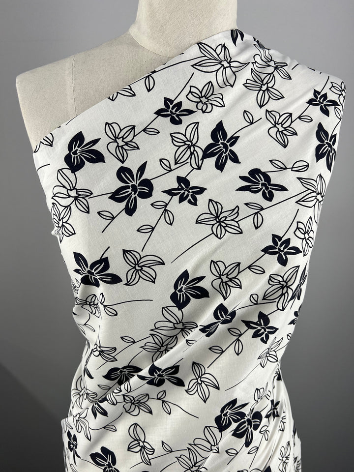 A fabric with a white background and a black floral pattern displayed on a mannequin. This Super Cheap Fabrics' Printed Linen - Mono Lotus - 150cm features various large and small flowers with branches and leaves, draped elegantly to showcase its design and texture, ideal for garment creations or even home decor projects.