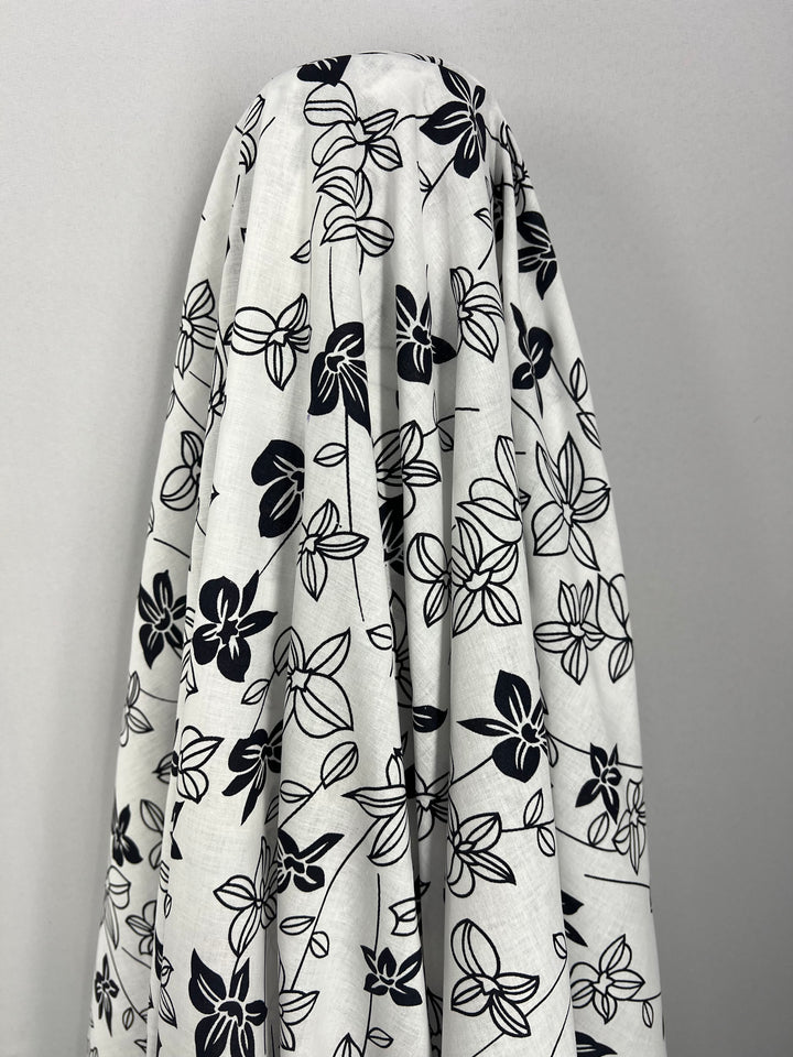 A draped piece of Super Cheap Fabrics' Printed Linen - Mono Lotus - 150cm with a black floral print. The fabric is arranged in soft, flowing folds against a neutral gray background. Perfect for home decor, the black flowers and leaves contrast sharply with the white, creating a striking visual pattern.