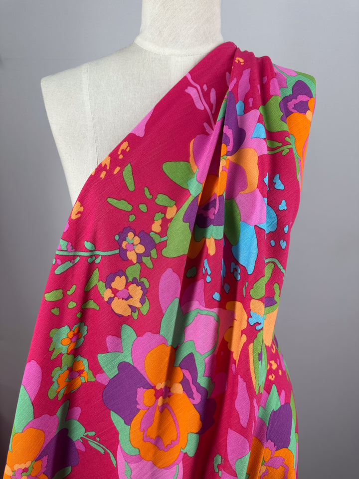 A mannequin displays Super Cheap Fabrics' Printed Linen Blend - Wild Fuchsia (145cm), featuring vivid floral patterns in orange, purple, green, and blue on bright pink—perfect inspiration for summer jackets against a plain gray background.