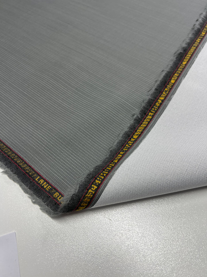 A close-up of a gray, slightly textured, medium weight fabric with a yellow and red trim along one edge, laying on a smooth, white surface. The edge of the Merino Wool Suiting - Gray & White Stripe - 155cm from Super Cheap Fabrics is slightly folded, revealing the white underside. The text on the trim is partially visible.