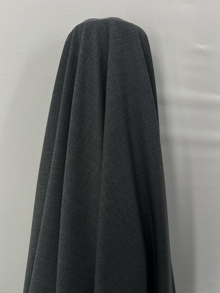 A dark gray Wool Suiting - Charcoal - 152cm from Super Cheap Fabrics is draped over an object, creating a smooth, flowing, and veiled appearance. The background is a plain white wall, contrasting with the luxurious texture of the dark fabric. The object underneath is completely obscured by the fabric.