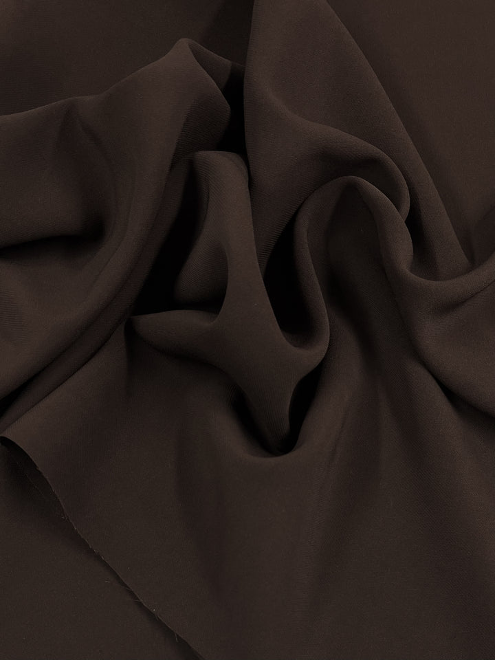 A close-up image of dark brown Premium Viscose Suiting - Choc Martini - 130cm with a soft, smooth texture by Super Cheap Fabrics. The fabric is loosely gathered, creating gentle folds and creases. The material appears to have a slight sheen, highlighting the depth and richness of the choc martini color.