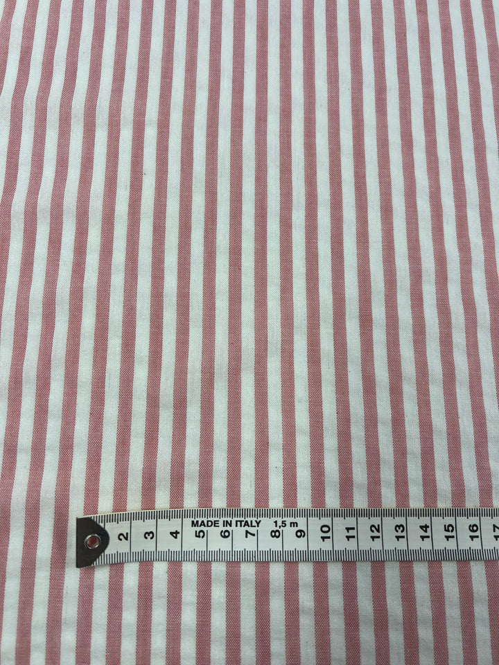 A piece of fabric with vertical pink and white stripes. A measuring tape, marked in both centimeters and inches, is placed horizontally at the bottom of the image, showing the fabric's stripes against the measurements. The breathable Linen Cotton - Pink Stripe - 145cm by Super Cheap Fabrics indicates it is made in Italy.